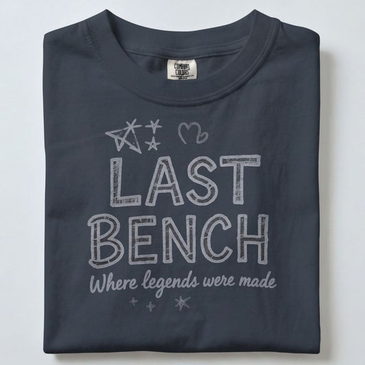 A folded dark gray t-shirt with a white graphic print that reads 'LAST BENCH Where legends were made' with stars and a heart, shown on a light gray background.