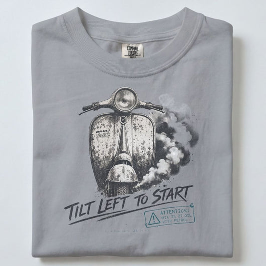 A folded light gray crewneck t-shirt featuring a black and white graphic of a vintage scooter with the text 'TILT LEFT TO START' and a small warning label graphic.