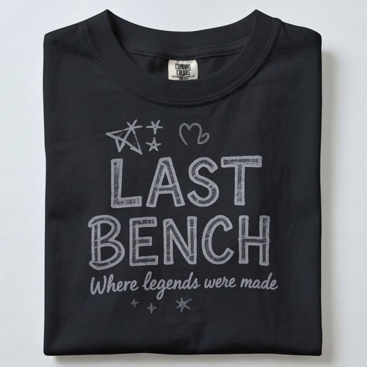A folded black t-shirt with a white graphic print that reads 'LAST BENCH Where legends were made' decorated with hand-drawn style stars and a heart, shown on a light gray background.