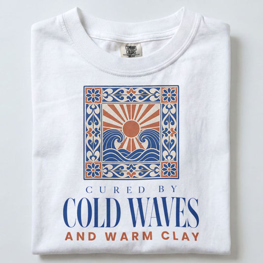 A folded white t-shirt with a graphic print featuring a stylized sun and waves inside a decorative border, with the text 'CURED BY COLD WAVES AND WARM CLAY' printed below.
