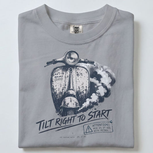 A folded light gray t-shirt featuring a black and white graphic of a vintage scooter with the text 'TILT RIGHT TO START' and a small warning label.