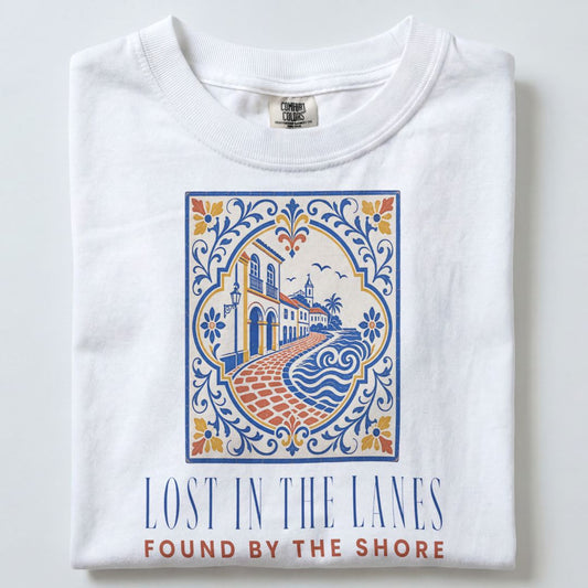 A folded white t-shirt with a colorful graphic print of a coastal town scene and the text 'LOST IN THE LANES FOUND BY THE SHORE' printed below it.